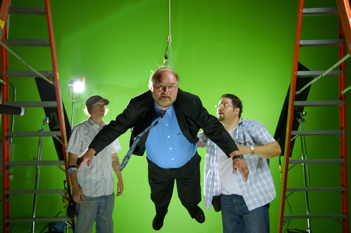 man hanging from a rope against a greenscreen in a bts shot of a film shoot