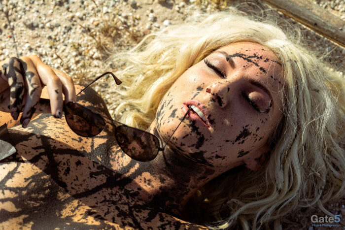 a blonde woman with black oil streaks on her lies on the desert floor in a fashion shot